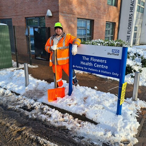 Council worker in high visibility jacket standing next to "The Flowers Health Care" NHS sign whilst clearing snow 