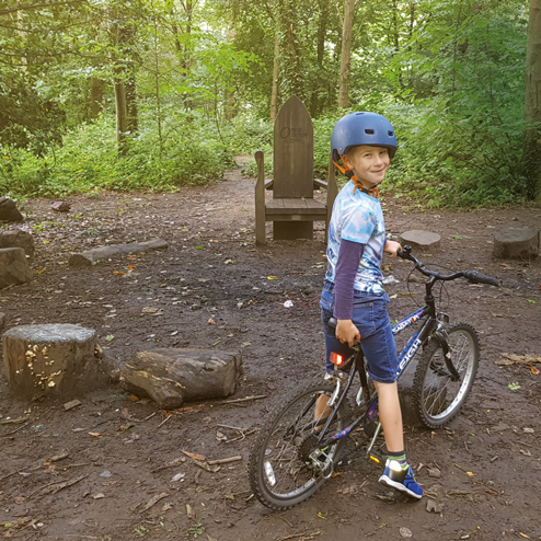 A boy sits on his bike looking back at the camera whilst wearing a helmet. A wooden throne sits in the background, whilst the photo is taken in a forest