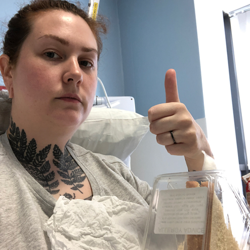 A lady takes a selfie in a hospital bed with her thumb up, and a sandwich on a table in front of her