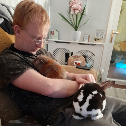 A man sits on a sofa with a guinea pig on his chest and a rabbit on his lap