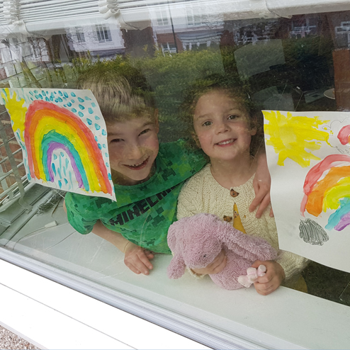 A boy and a girl behind window which has 2 painted rainbows on paper stuck to it