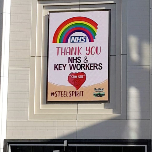 Sign  on building with rainbow arching over NHS logo, and text below reading "THANK YOU NHS & KEY WORKERS". A red love heart with "STAY SAFE" and a smiley face in it. At the bottom, it reads #STEELSPIRIT, which is beside the Sheffield City Council logo