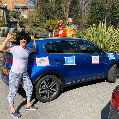 A lady is stood, looking happy, in front of blue car which has 4 rainbows stuck on it. A dog is stood on top of the car, and 2 people are behind the car in fancy dress.