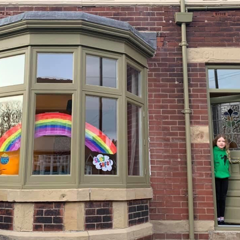 A girl stands in a doorway next to bay windows which have a rainbow reaching across the middle 3 of 5