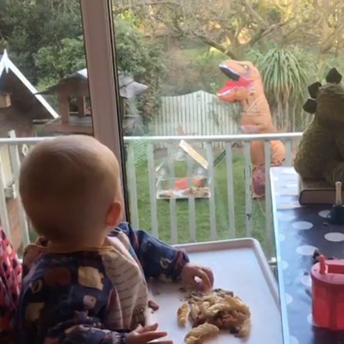 A toddler looking into his garden where a person is mowing the lawn whilst in a dinosaur fancy dress costume