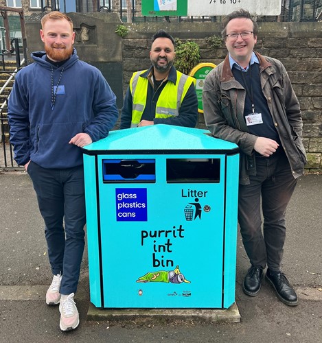 Three men - artist Luke Horton, Khalid Mahmood of Imperial Signs and Cllr Joe Otten - stand next to a blue painted bin in front of a school