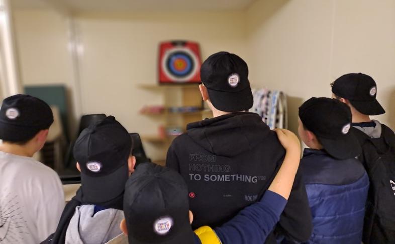 Young people playing darts. They are facing the dartboard and are all wearing baseball caps