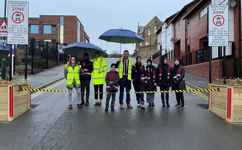 image of pupils and adults stood in the road in front of black and yellow tape. Two adults are holding umbrellas