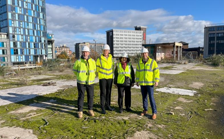 4 people in hi viz jackets and white hard hats stand at the Castle site with high rise buildings in the background