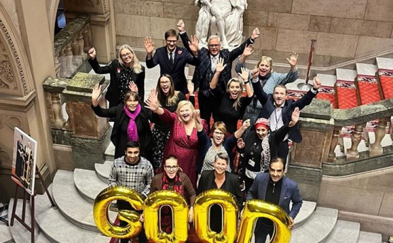 A group of people stand at the bottom of a large staircase with a statue behind them. The front row of people are holding up golden coloured balloon letters spelling the word gold in capital letters. They are all cheering 