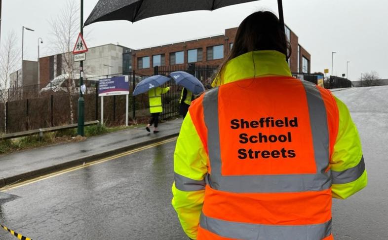 Person stood on the street holding an umbrella. It is raining. They are wearing a hi-viz jacket in orange and yellow with the words Sheffield School Streets printed on the back 