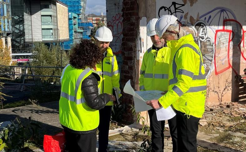 Group of people wearing hi viz jackets and hard hats studying a map. They are on a work site with demolished buildings. There is some graffiti on the wall behind them 