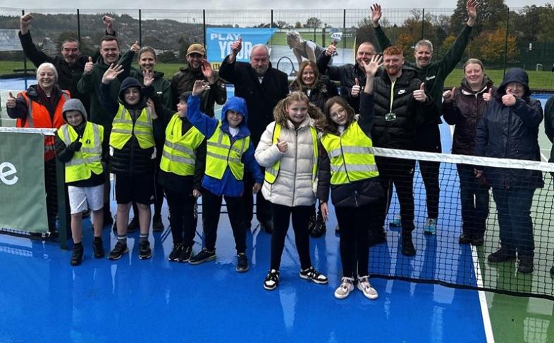 A group of adults and children cheering in an outdoor tennis court. The children are wearing hi viz jackets 