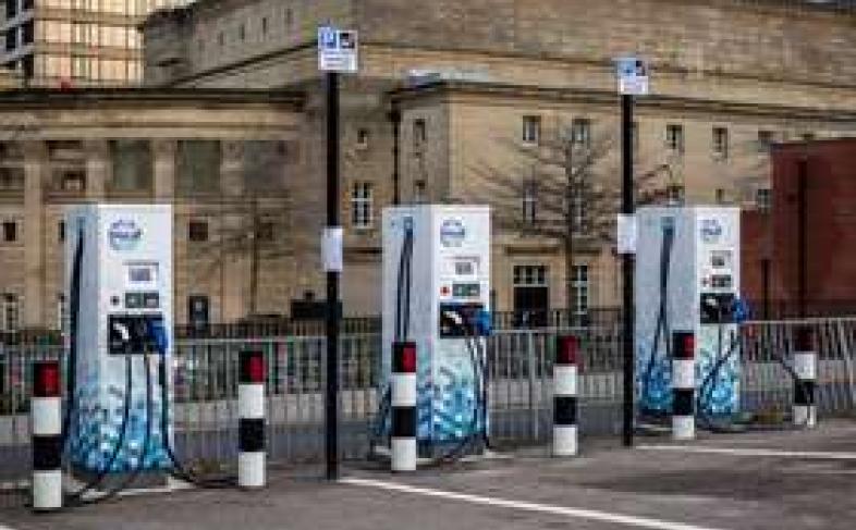 Three blue and white electric charging points stand in a car park in painted parking bays in front of a civic building on a dull, cloudy day