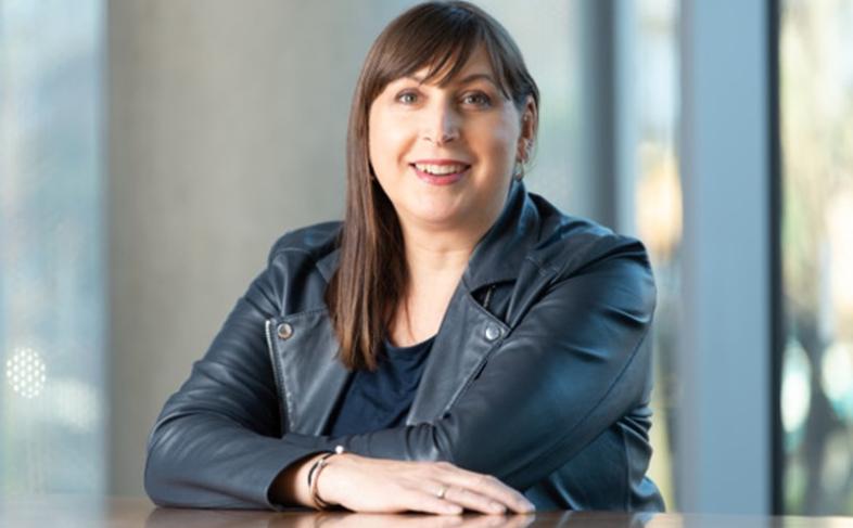 Alex Willard smiles at the camera while sat at a table in a corporate looking office, wearing a black leather jacket