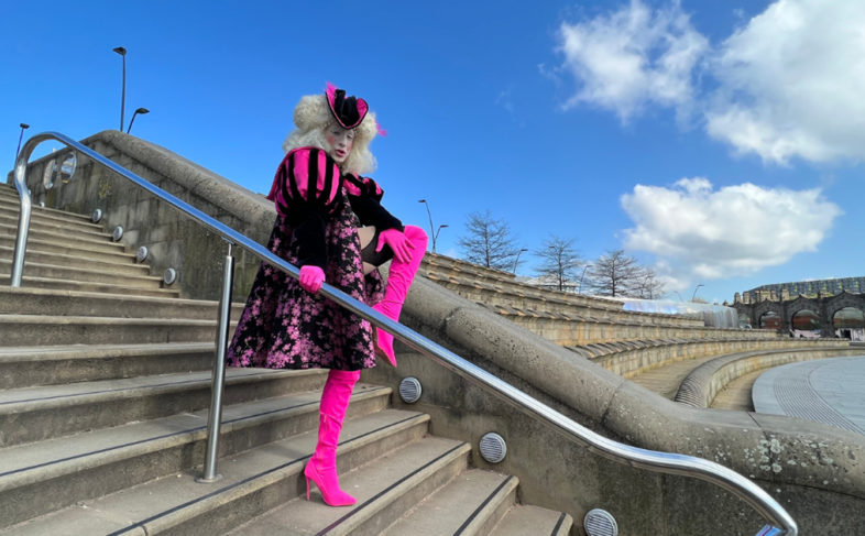 A drag artist dressed in flamboyant clothing prepares to do a high kick while standing on a set of stairs with a metal hand rail going down the middle