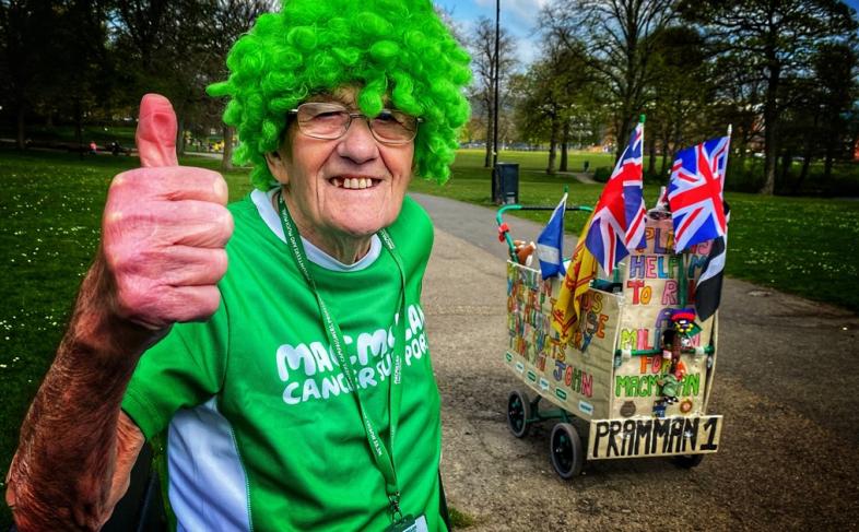 John Burkill with a green curly wig and green Macmillian t shirt. His pram is in the background and he's holding his thumb up and smiling. 