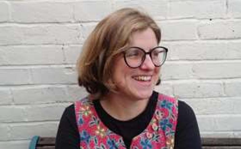 Mandy Fletcher smiles while looking to the side, wearing black-rimmed glasses and a pink, flowery top. She is sat on a bench in front of a white brick wall