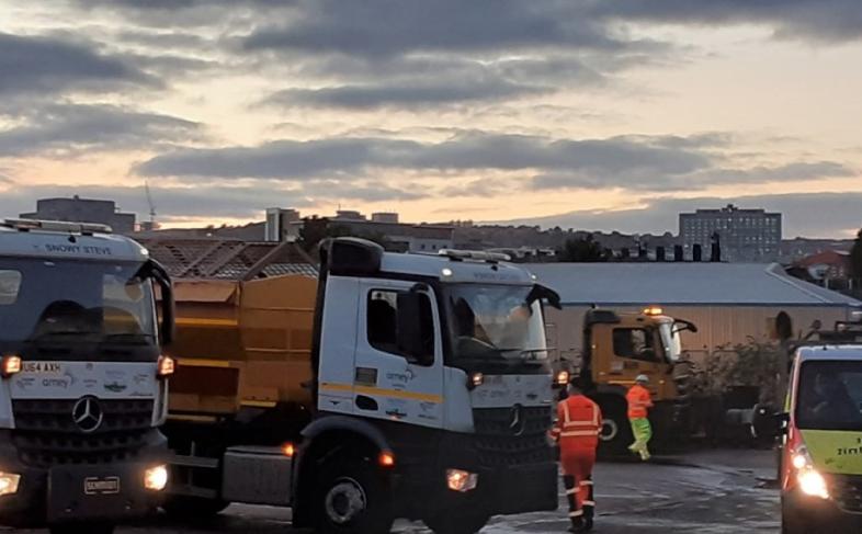 Two gritter lorries stand on the right hand side of the picture with two workmen in front of them and a works van on the right hand side