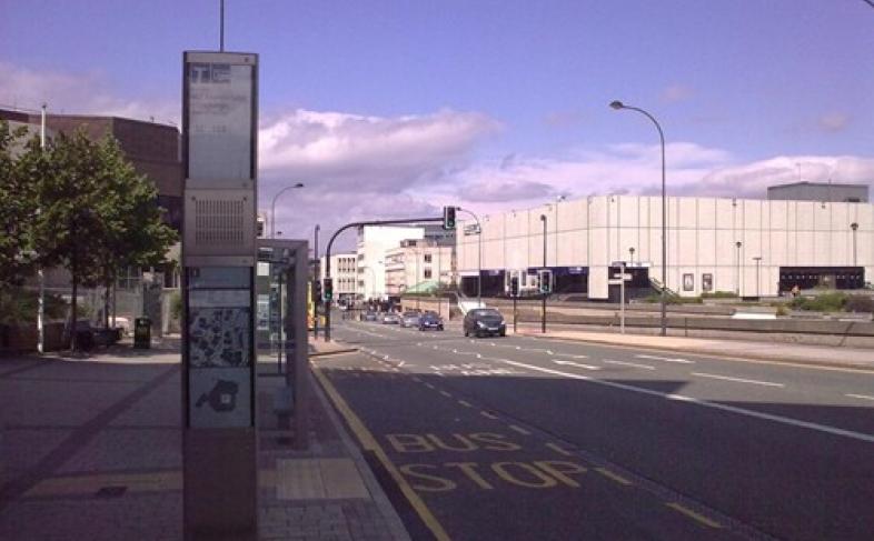 An empty Sheffield street with just a single car travelling towards the camera, the road has buildings and trees on either side with a bus stop in the foreground