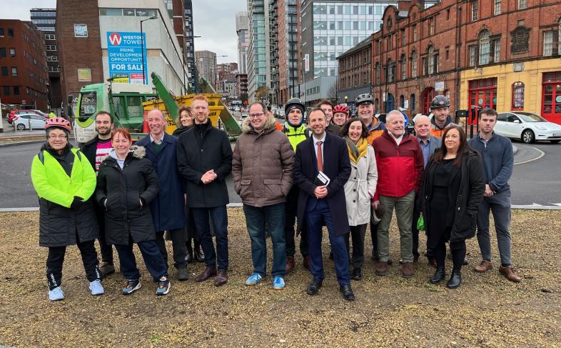 A group of people stand in front of the new West Bar roundabout in Sheffield City Centre with tall buildings on either side of the picture.