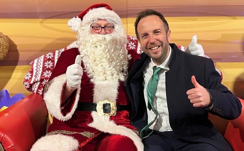 Cllr Ben Miskell sits next to Santa Claus, both have their thumbs up and they are in Santa's grotto