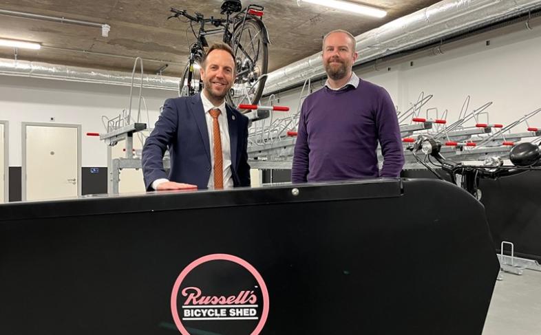 Cllr Ben Miskell and Russell Cutts stand behind a black board with 'Russell's Bicycle Shed' written on the front, they are both standing in a cycle hub with bikes on ranks above them