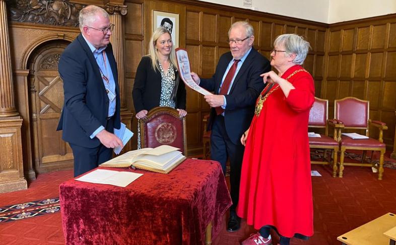 Two women and two men stand in one of the Town Hall rooms, over a table with a book and red table cloth on it