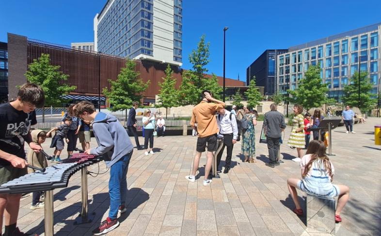 Several people can be seen playing with musical instruments with a road in the middle ground and buildings in the back ground