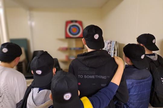 Young people playing darts. They are facing the dartboard and are all wearing baseball caps