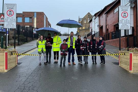 image of pupils and adults stood in the road in front of black and yellow tape. Two adults are holding umbrellas