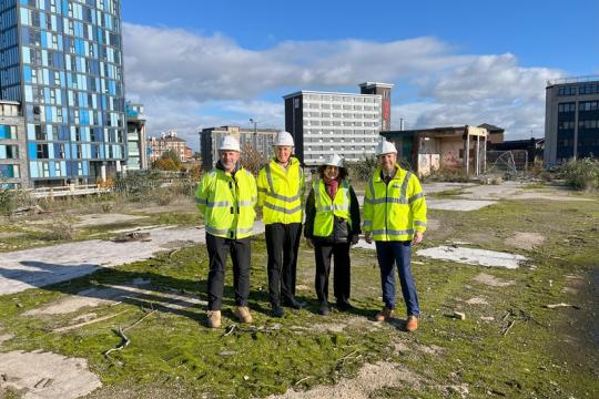 4 people in hi viz jackets and white hard hats stand at the Castle site with high rise buildings in the background