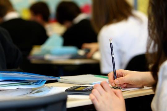 Classroom with a closeup of a students desks and hands holding and writing with a pencil - classmates blurred in the background