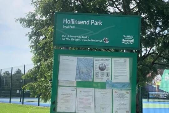 A green sign of Hollinsend Park with notices underneath, trees and playing pitched in the background