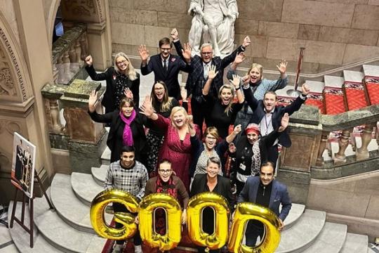 A group of people stand at the bottom of a large staircase with a statue behind them. The front row of people are holding up golden coloured balloon letters spelling the word gold in capital letters. They are all cheering 