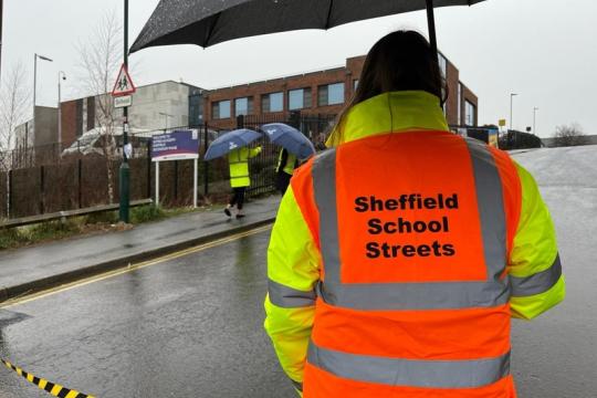 Person stood on the street holding an umbrella. It is raining. They are wearing a hi-viz jacket in orange and yellow with the words Sheffield School Streets printed on the back 