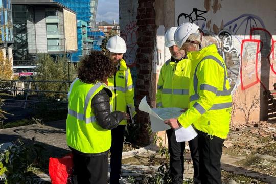 Group of people wearing hi viz jackets and hard hats studying a map. They are on a work site with demolished buildings. There is some graffiti on the wall behind them 