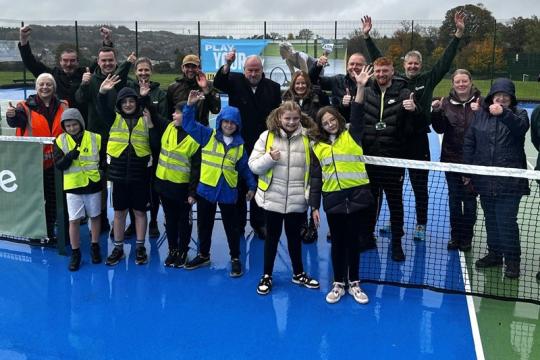 A group of adults and children cheering in an outdoor tennis court. The children are wearing hi viz jackets 