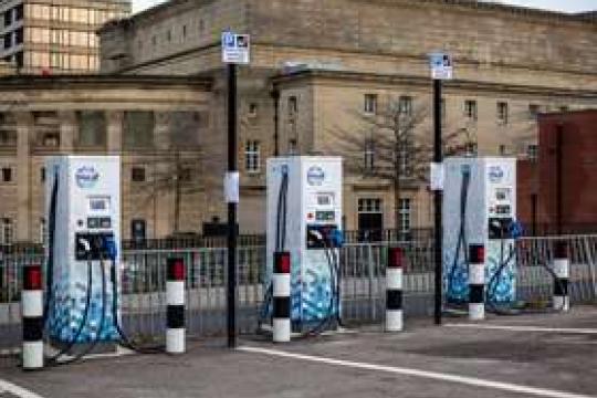 Three blue and white electric charging points stand in a car park in painted parking bays in front of a civic building on a dull, cloudy day