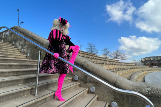 A drag artist dressed in flamboyant clothing prepares to do a high kick while standing on a set of stairs with a metal hand rail going down the middle