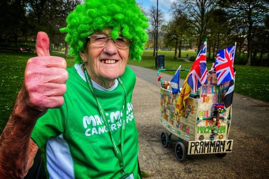 John Burkill with a green curly wig and green Macmillian t shirt. His pram is in the background and he's holding his thumb up and smiling. 