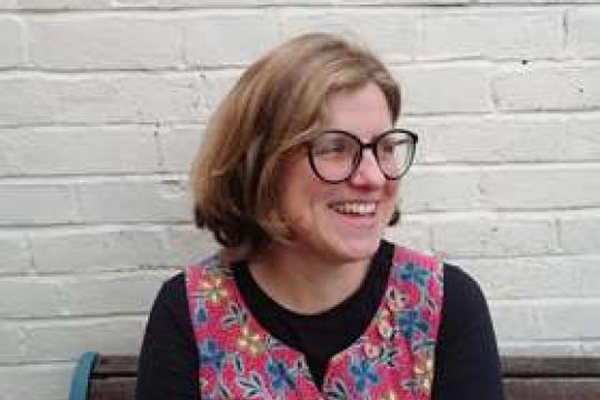 Mandy Fletcher smiles while looking to the side. She is sat on a bench in front of a white brick wall