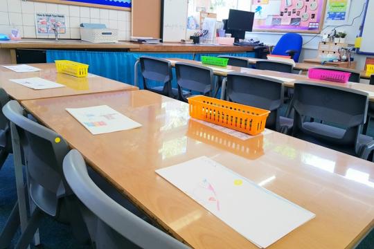 An empty school classroom with a close up view of student's tables and chairs with paper on top