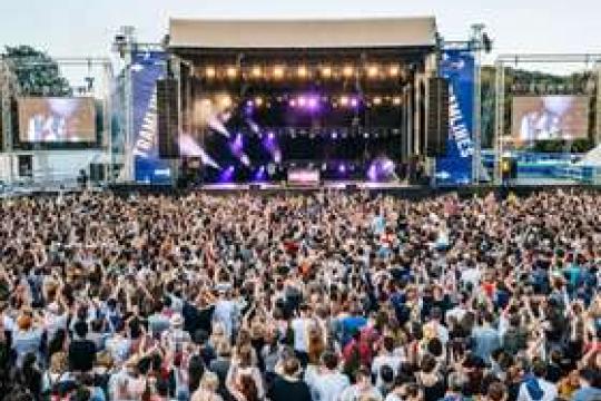 A packed crowd at Tramlines watch a performer under a cloudy sky as the stage lights shine brightly