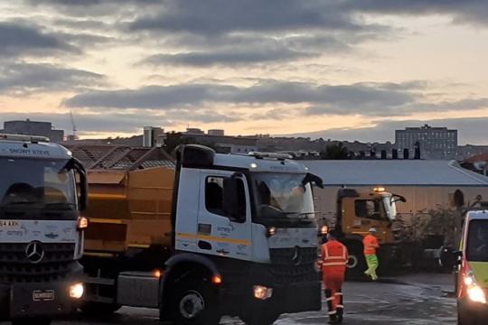 Two gritter lorries stand on the right hand side of the picture with two workmen in front of them and a works van on the right hand side