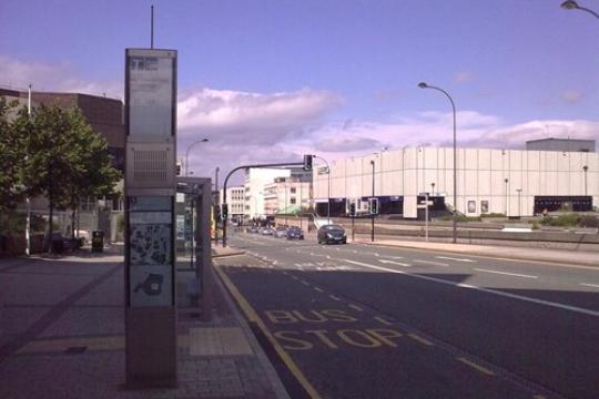 An empty Sheffield street with just a single car travelling towards the camera, the road has buildings and trees on either side with a bus stop in the foreground