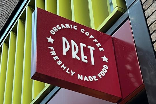 A Pret sign hanging from the side of the new store in Sheffield city centre, the image shows the sign with a yellow, metal, design of the building in the background.