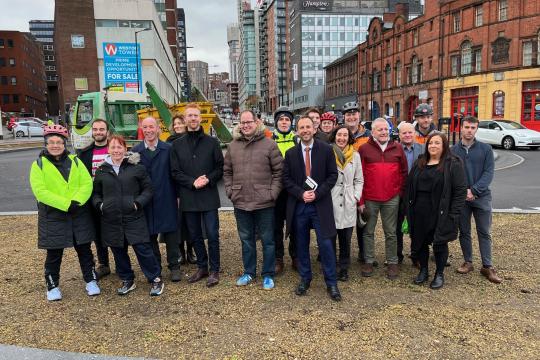 A group of people stand in front of the new West Bar roundabout in Sheffield City Centre with tall buildings on either side of the picture.