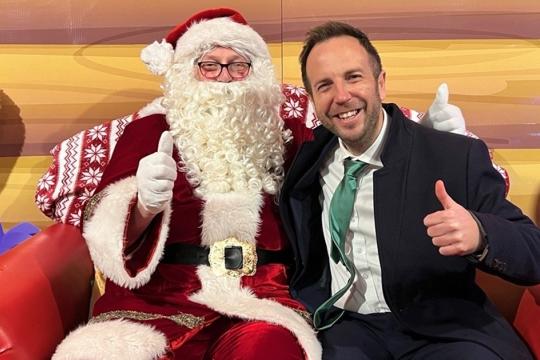 Cllr Ben Miskell sits next to Santa Claus, both have their thumbs up and they are in Santa's grotto