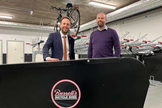 Cllr Ben Miskell and Russell Cutts stand behind a black board with 'Russell's Bicycle Shed' written on the front, they are both standing in a cycle hub with bikes on ranks above them